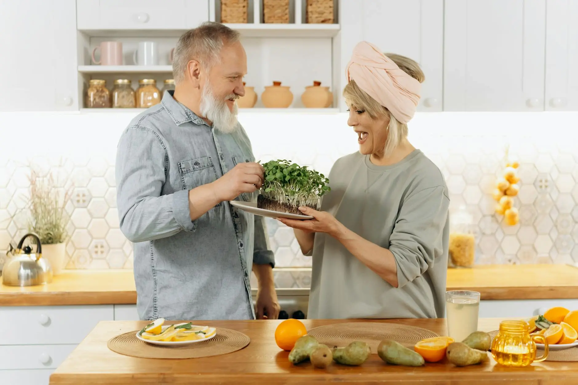 An older couple in a kitchen, smiling at each other. The man, wearing a denim shirt, is holding a plate with microgreens, while the woman in a headwrap and sweater presents it to him. The countertop has a plate of sliced fruit, whole fruit, and a jar of juice. The background features kitchen cabinets and decor.